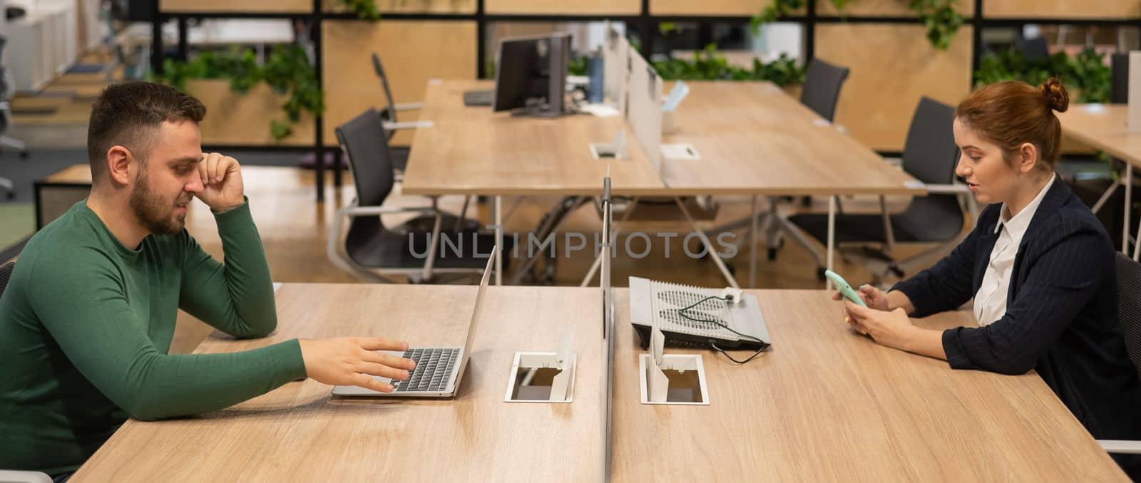 Red-haired caucasian woman uses a smartphone and a bearded man works behind a laptop in an open space office