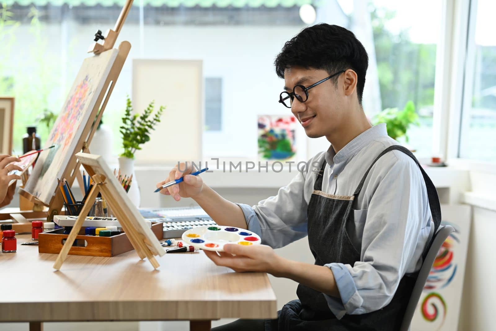 Smiling asian man student in apron painting in watercolor on easel. Art, creative hobby and leisure activity concept.