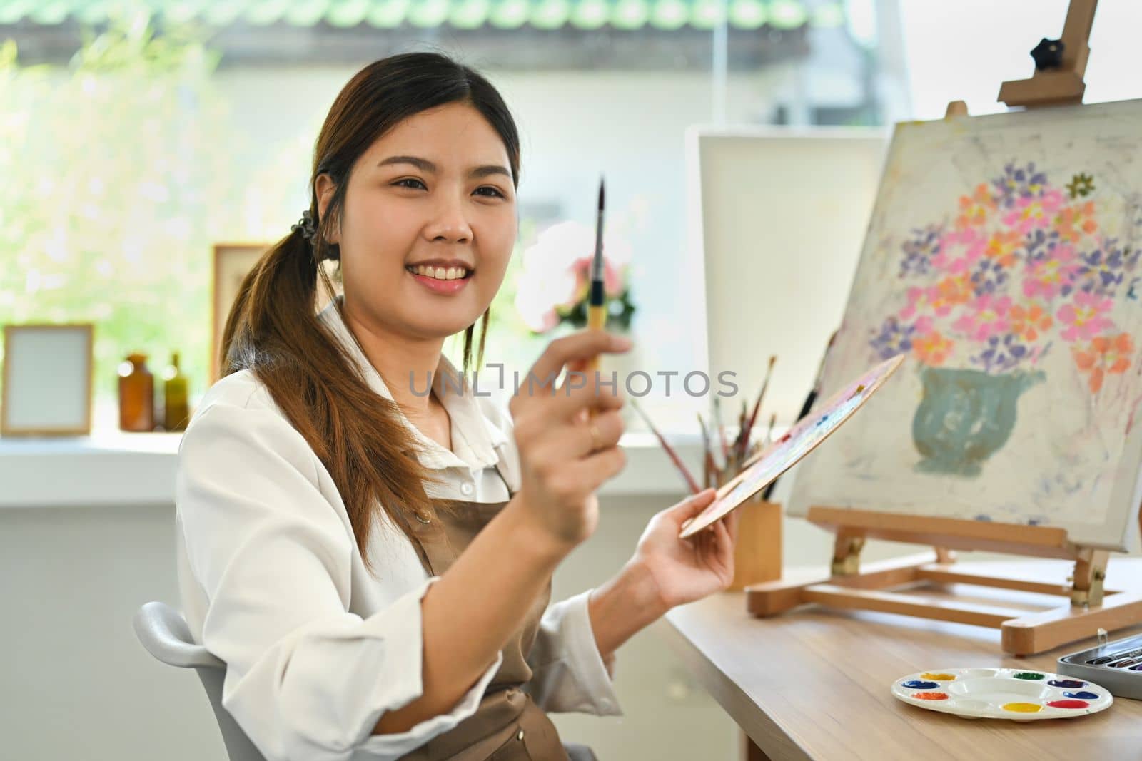 Pretty young woman artist holding paintbrush and palette, sitting front of easel in art studio.