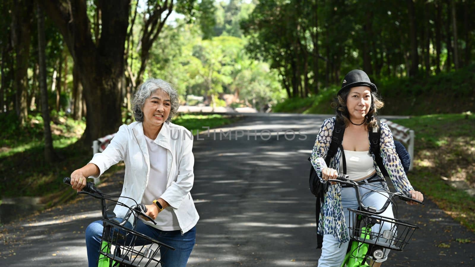 Two smiling middle aged women cycling in nature park on beautiful day. Retirement people lifestyle, outdoor activity concept.