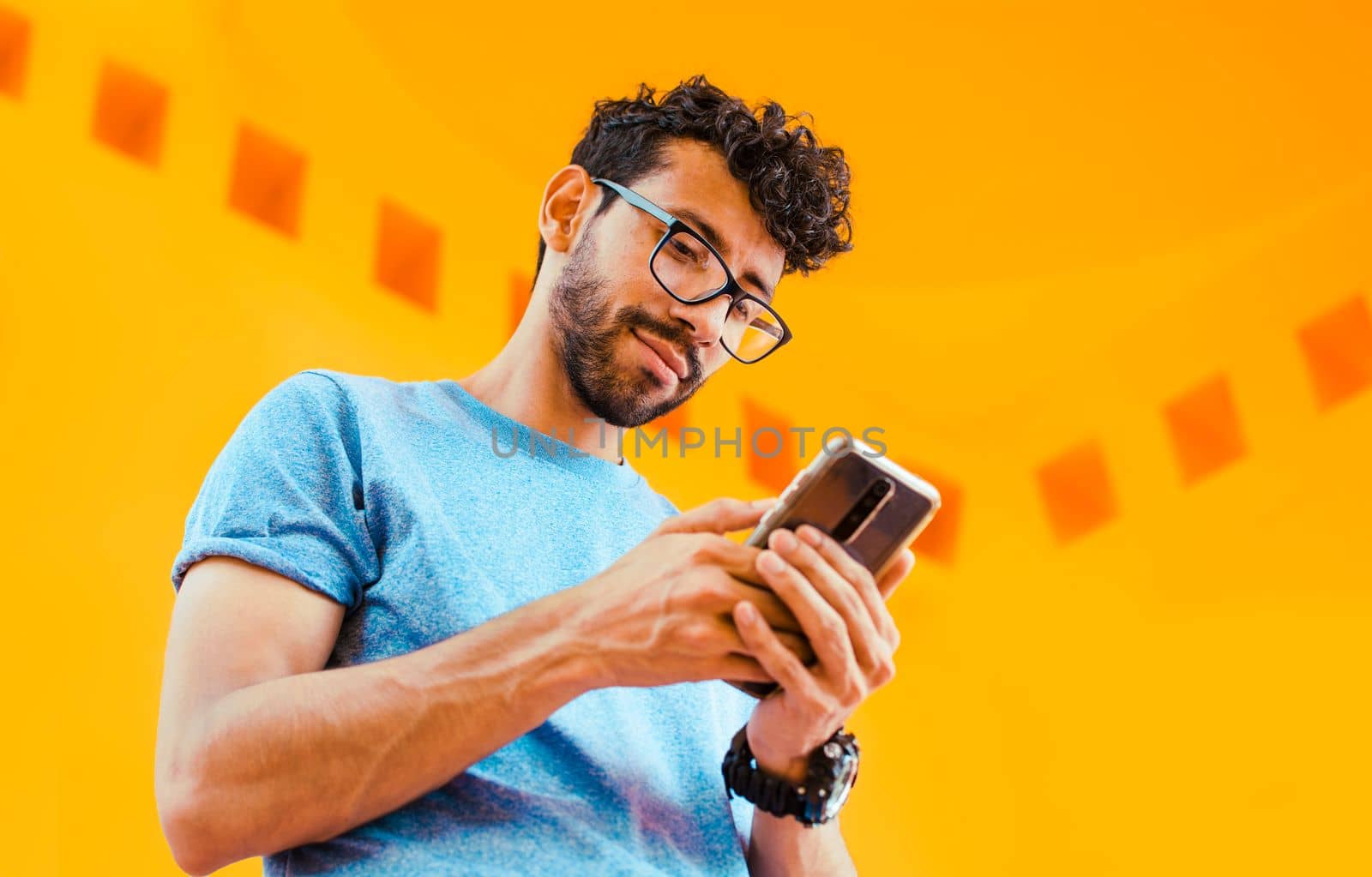 Low angle view of young man using cell phone outdoors. Guy sending a message with a cell phone near a yellow wall outdoors. Lifestyle of handsome bearded man using cell phone outdoors