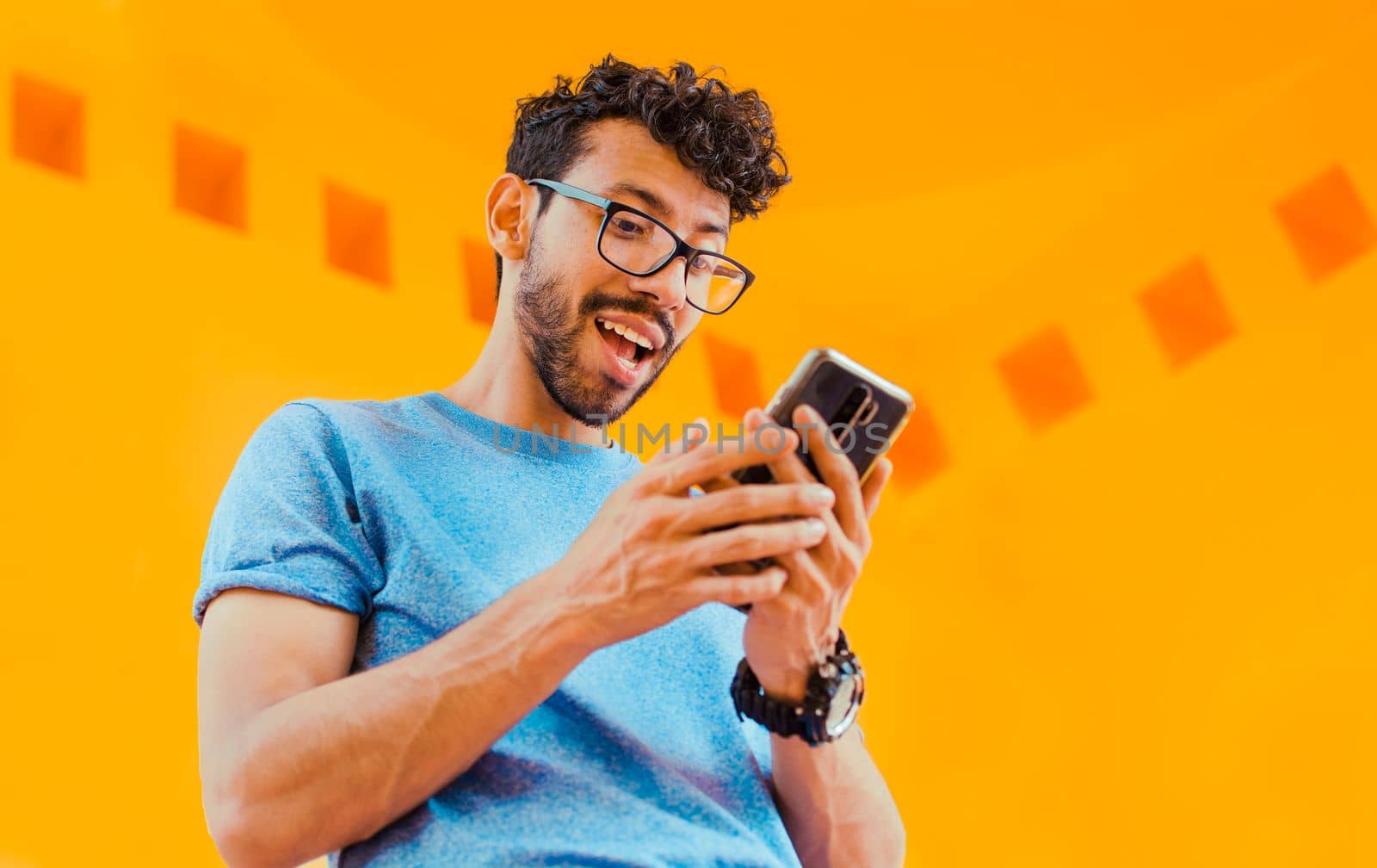 Happy guy sending a message with a cell phone near a yellow wall outdoors. Lifestyle of handsome bearded man using cell phone outdoors, Low angle view of young man using cell phone outdoors by isaiphoto
