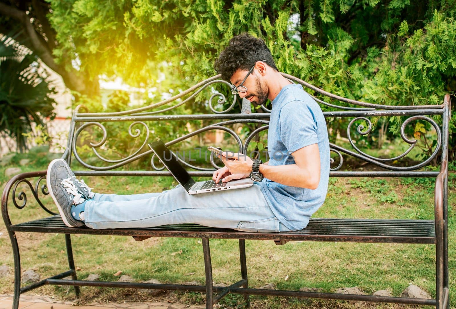 Freelancer man sitting in a park using laptop and cellphone. Man in a park working online with laptop. Relaxed man working with laptop outdoor, Young man in a park with laptop and cell phone