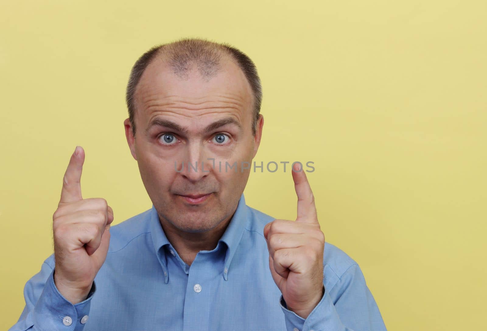 A man 45-55 years old in a blue shirt on a yellow background raised two hands with an index finger.