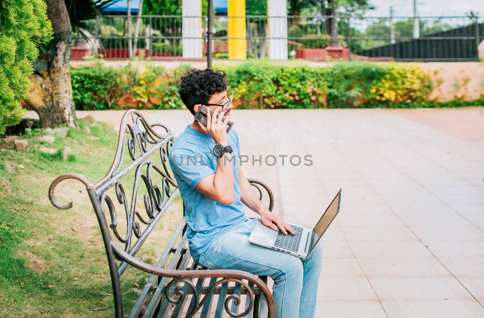 Young man sitting in a park working with his laptop and cell phone. Man in a park working online with laptop and calling on cellphone. Freelancer man working with laptop while calling on the phone