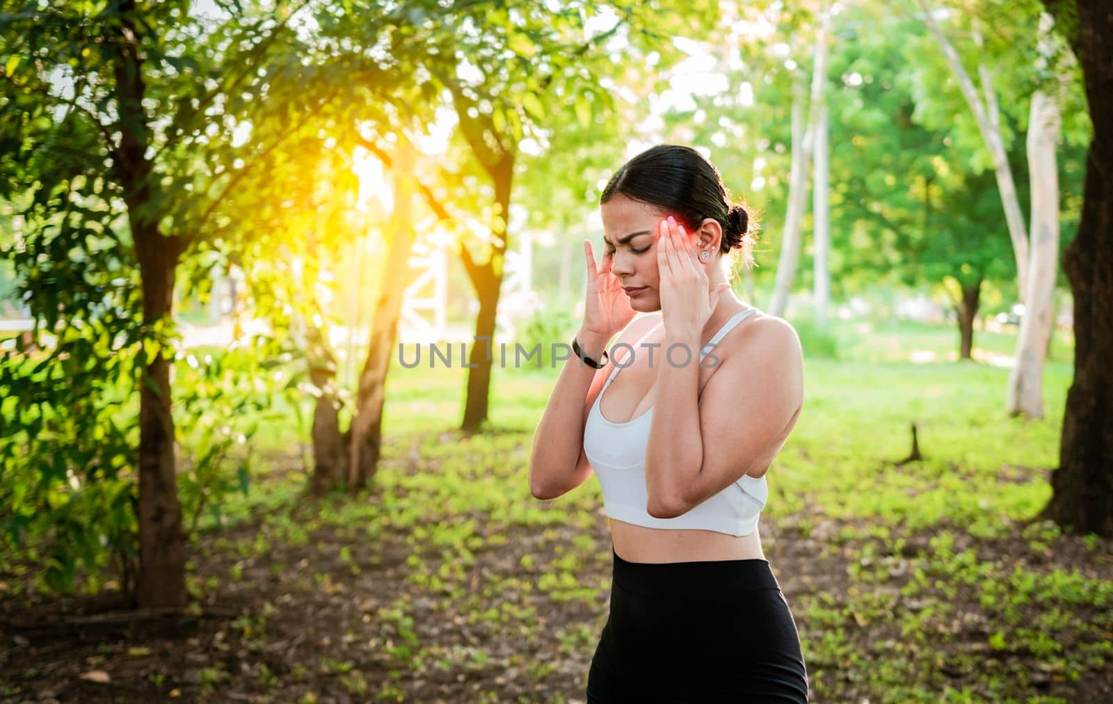Athlete girl with migraine in a park. Young female runner rubbing her head with migraine in a park. Runner woman with headache and fatigue in the park, Runner woman with headache in a park