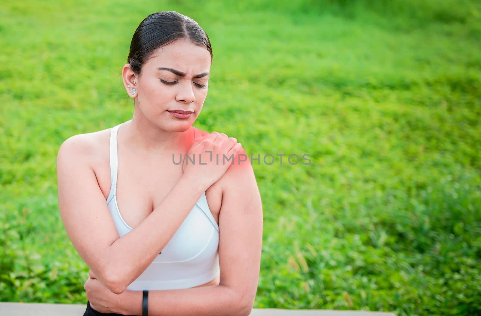 Young woman with shoulder pain sitting outside. Girl suffering from shoulder pain sitting outdoors. Woman sitting and stressed with shoulder pain outdoors. Girl with shoulder pain outdoors