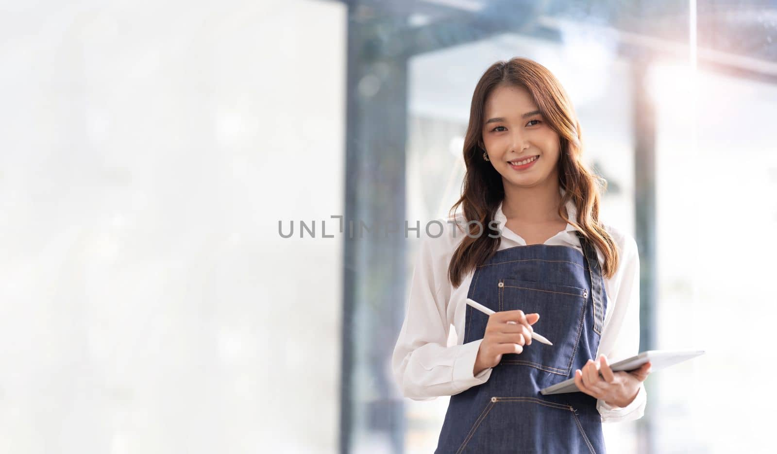 Beautiful asian young barista woman in apron holding tablet and standing in front of the door of cafe with open sign board. Business owner startup concept...