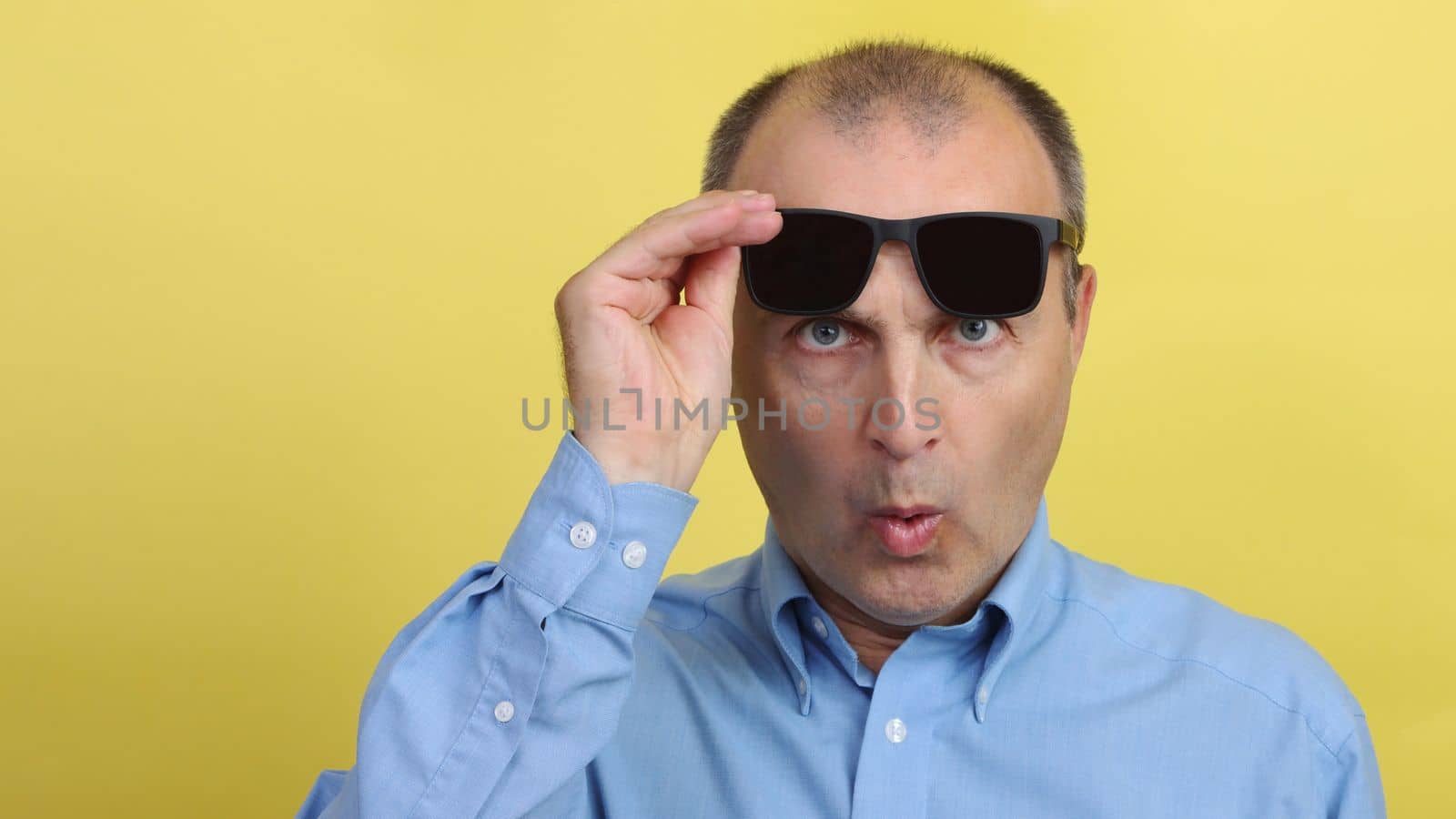 A man with glasses on a yellow background. Handsome surprised man in a blue shirt on a yellow background holds black glasses in his hands and looks at the camera.