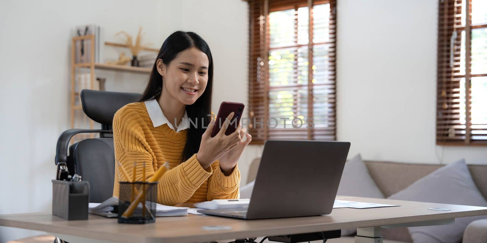 Smiling beautiful Asian businesswoman analyzing chart and graph showing changes on the market and holding smartphone at home by wichayada