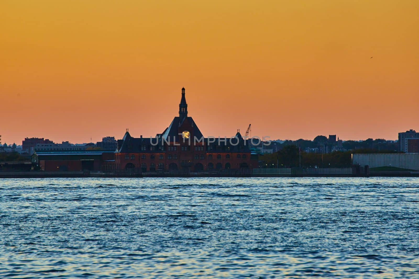 Image of New Jersey ferry station to Ellis Island from waters with glowing clock and orange dusk sky