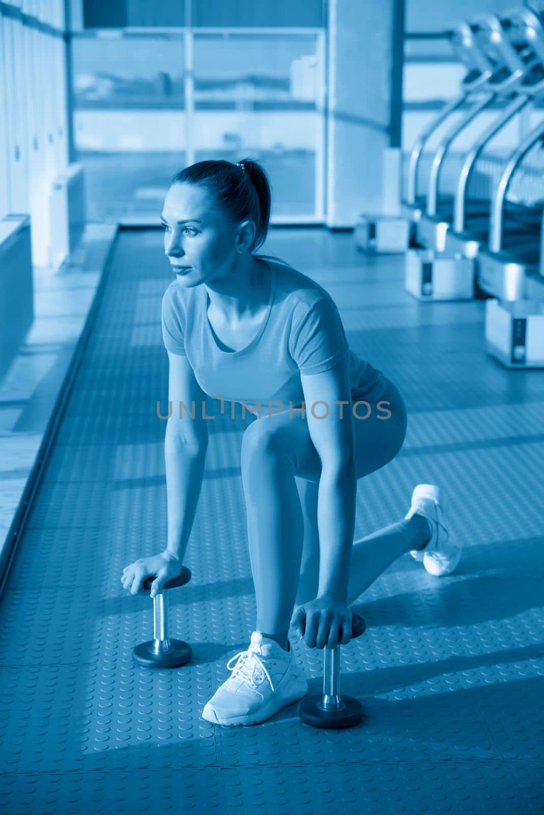Physically fit woman at the gym lifting dumbbells to strengthen her arms and biceps. Muscular woman sitting on exercise mat looking at her arms.