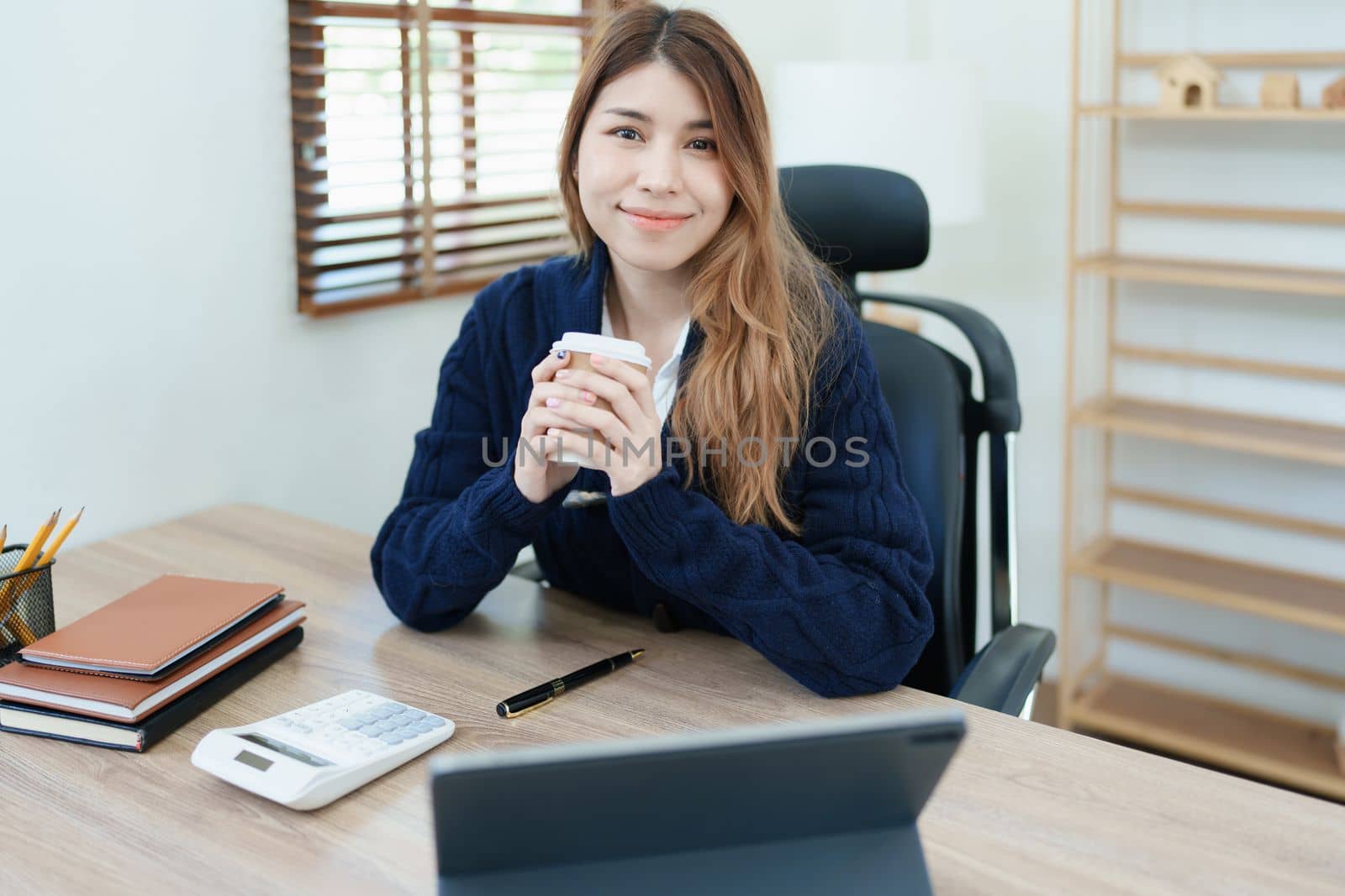 Portrait of an Asian business woman drinking coffee while working with a computer and financial statements documents on her desk.