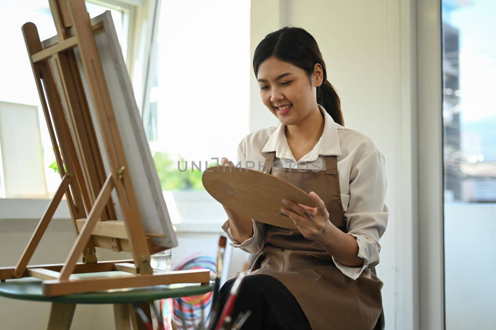 Smiling asian student woman in apron painting in watercolor on easel. Education, hobby, art concept by prathanchorruangsak