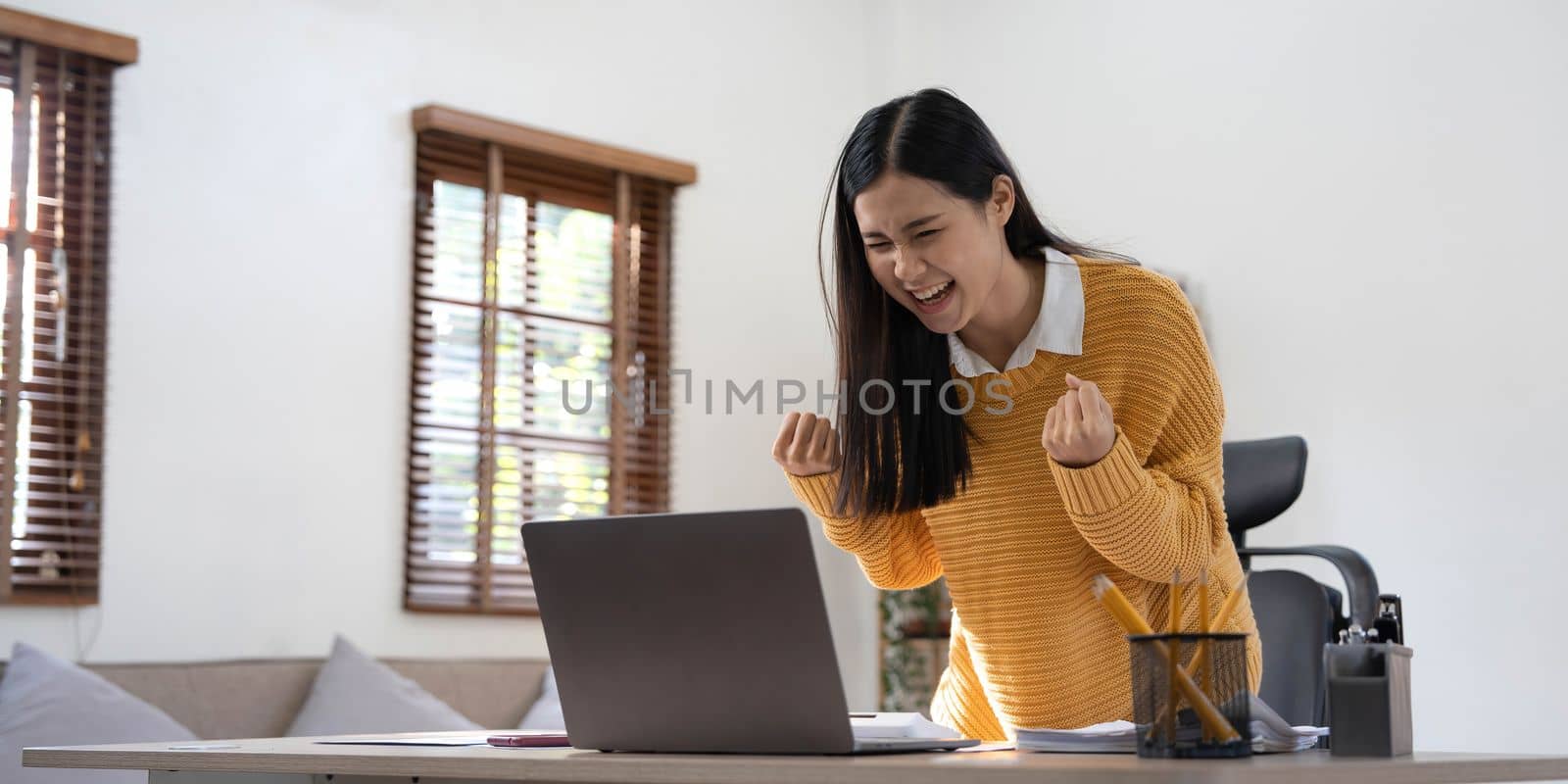 Excited happy woman looking at the laptop computer screen, celebrating an online win, overjoyed young asian female screaming with joy at home by wichayada