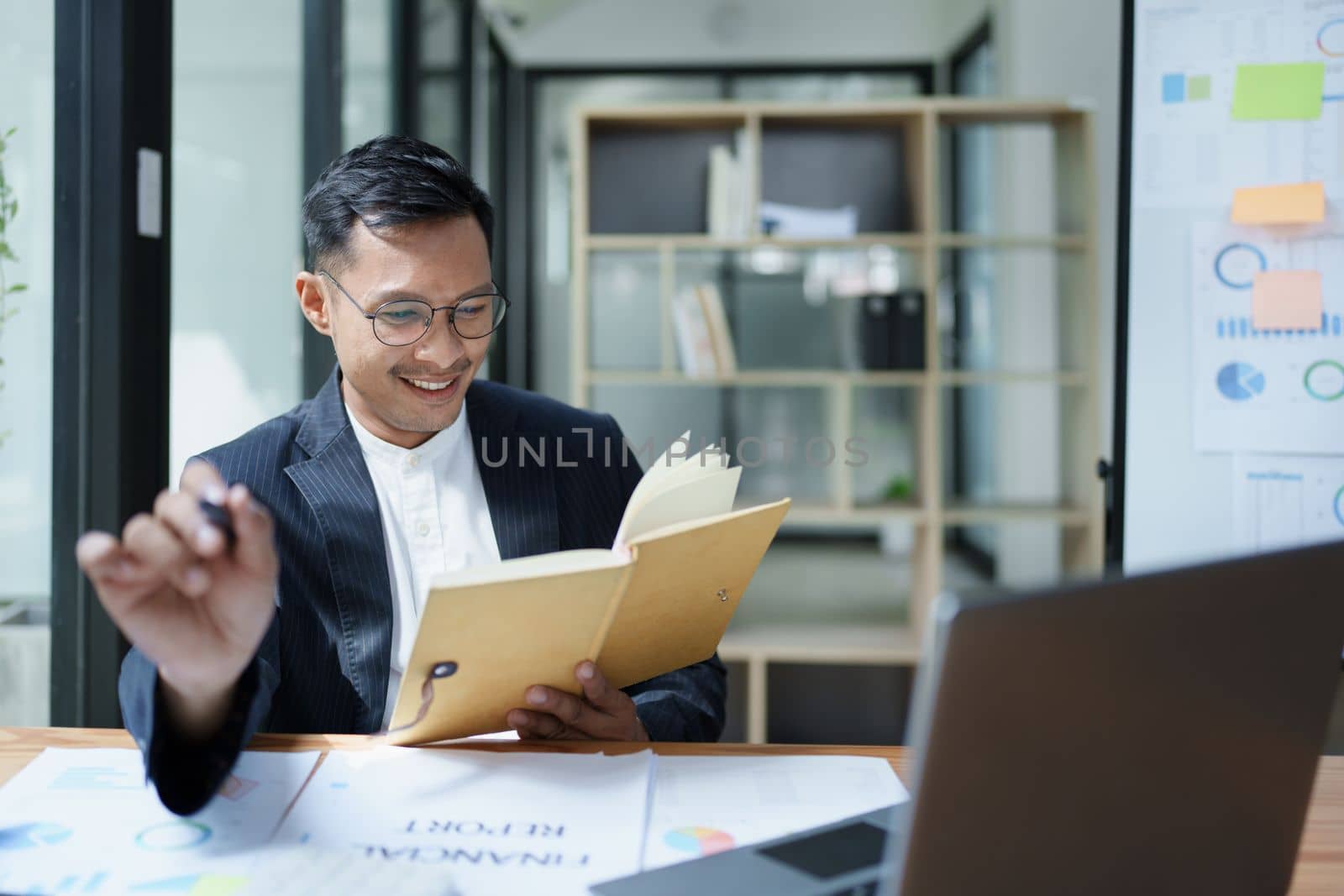 Portrait of a young Asian man showing a smiling face as she uses his notebook, computer and financial documents on her desk in the early morning hours.