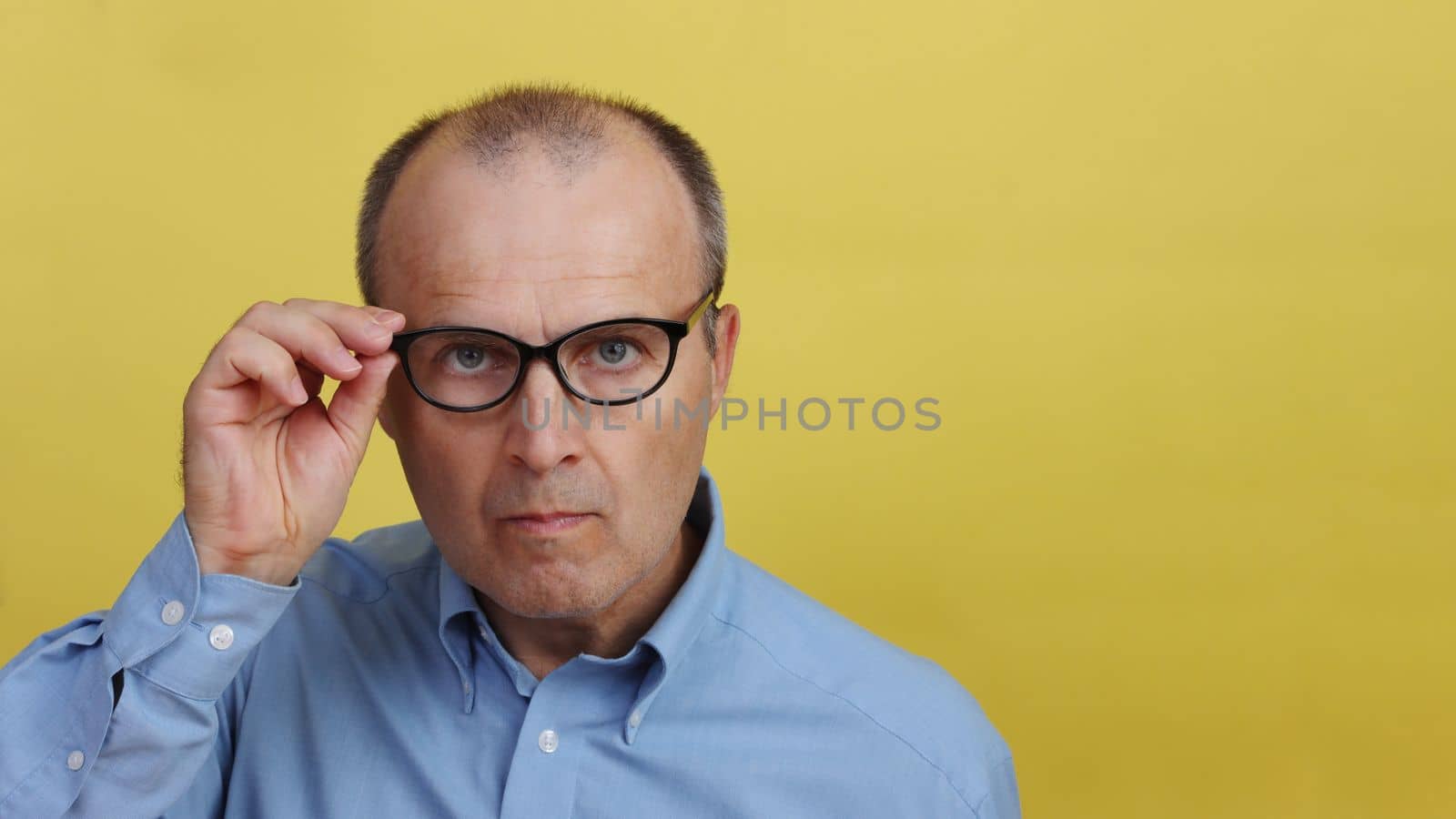 Handsome, intelligent-looking man, 45-55 years old, wearing a blue shirt against a yellow background, holding his glasses with his hand.