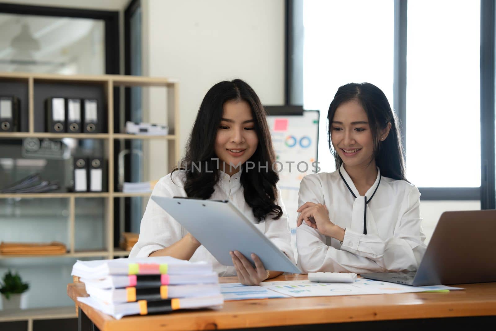 Two young Asian businesswomen show joyful expression of success at work smiling happily with a laptop computer in a modern office. by wichayada