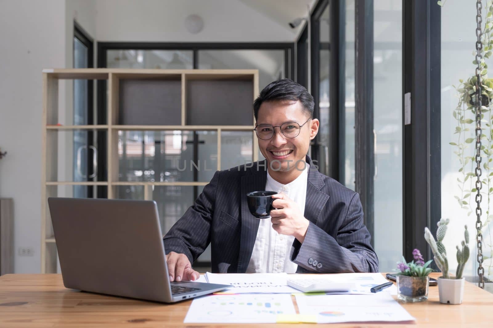 Young Asian businessman in suit and holding hot coffee cup and looking on laptop computer in office. Man in suit using laptop in well-lit workplace...