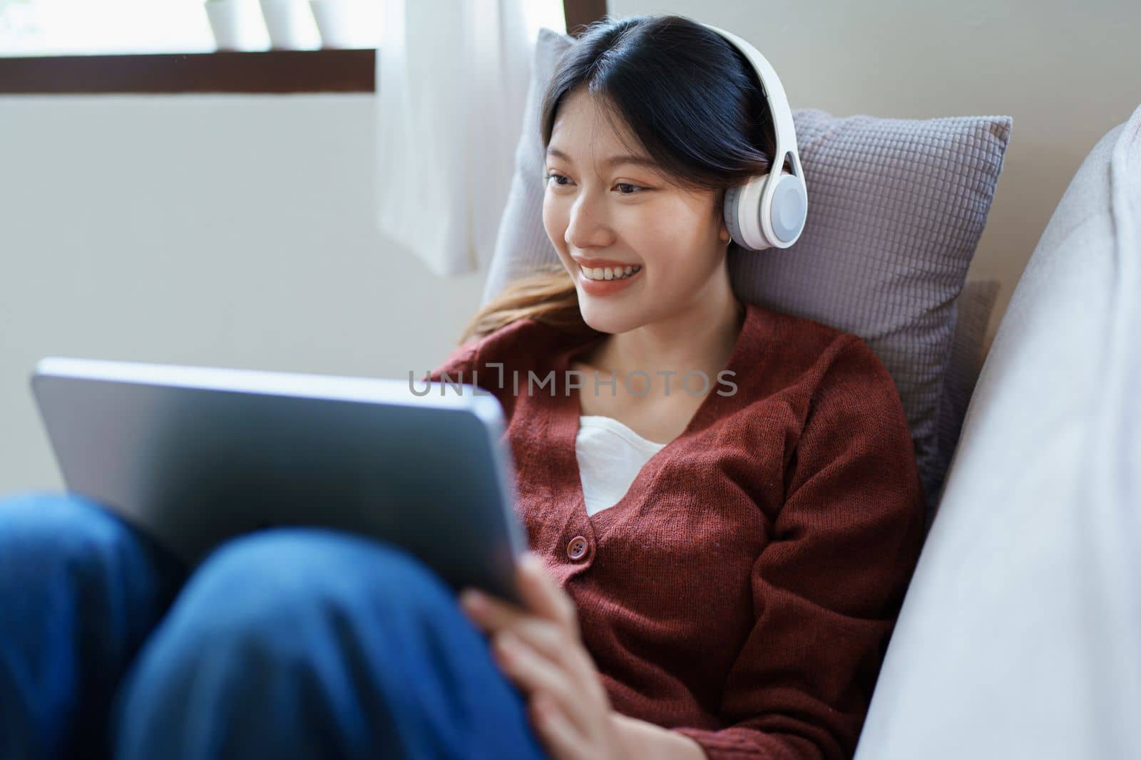 Portrait of asian woman using tablet and headphones relaxing on sofa at home by Manastrong