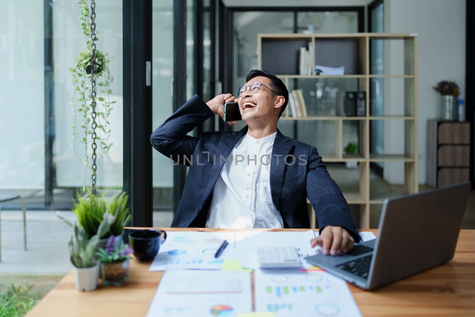 Portrait of a young Asian man showing a smiling face as she uses his phone, computer and financial documents on her desk in the early morning hours.