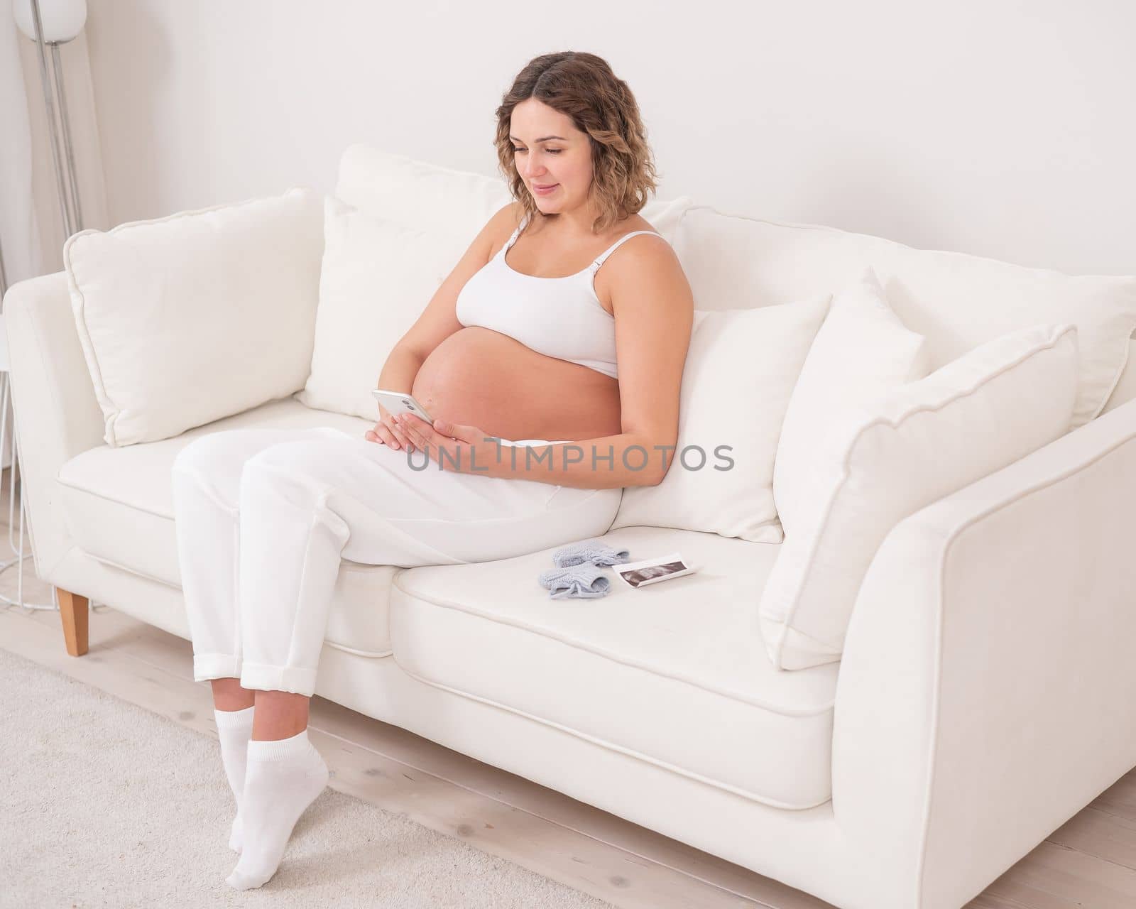 Pregnant woman sits on a white sofa and holds a smartphone. Fetal ultrasonic screening