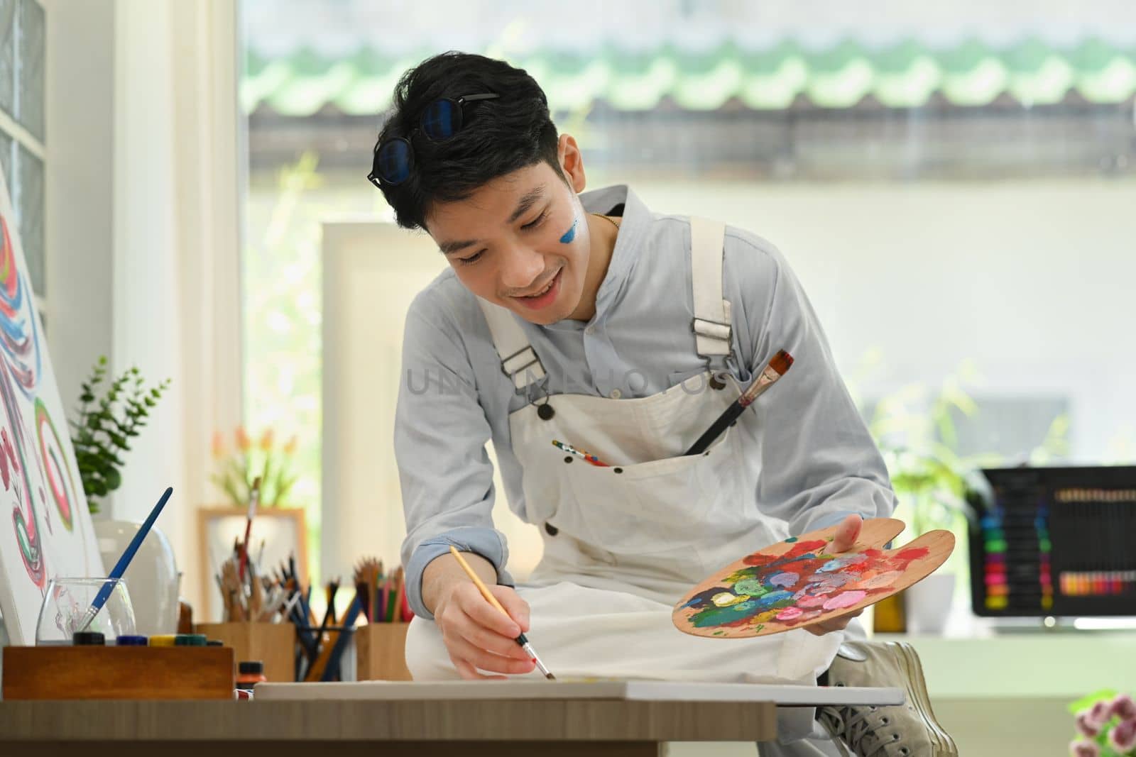 Happy asian male artist sitting in bright art workshop and enjoy creating his artwork.