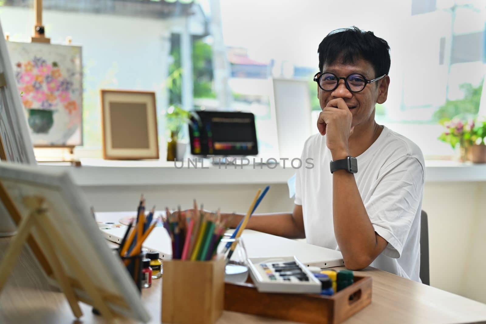 Happy asian man sitting in front of canvas in art studio and smiling to camera. Art and leisure activity concept.
