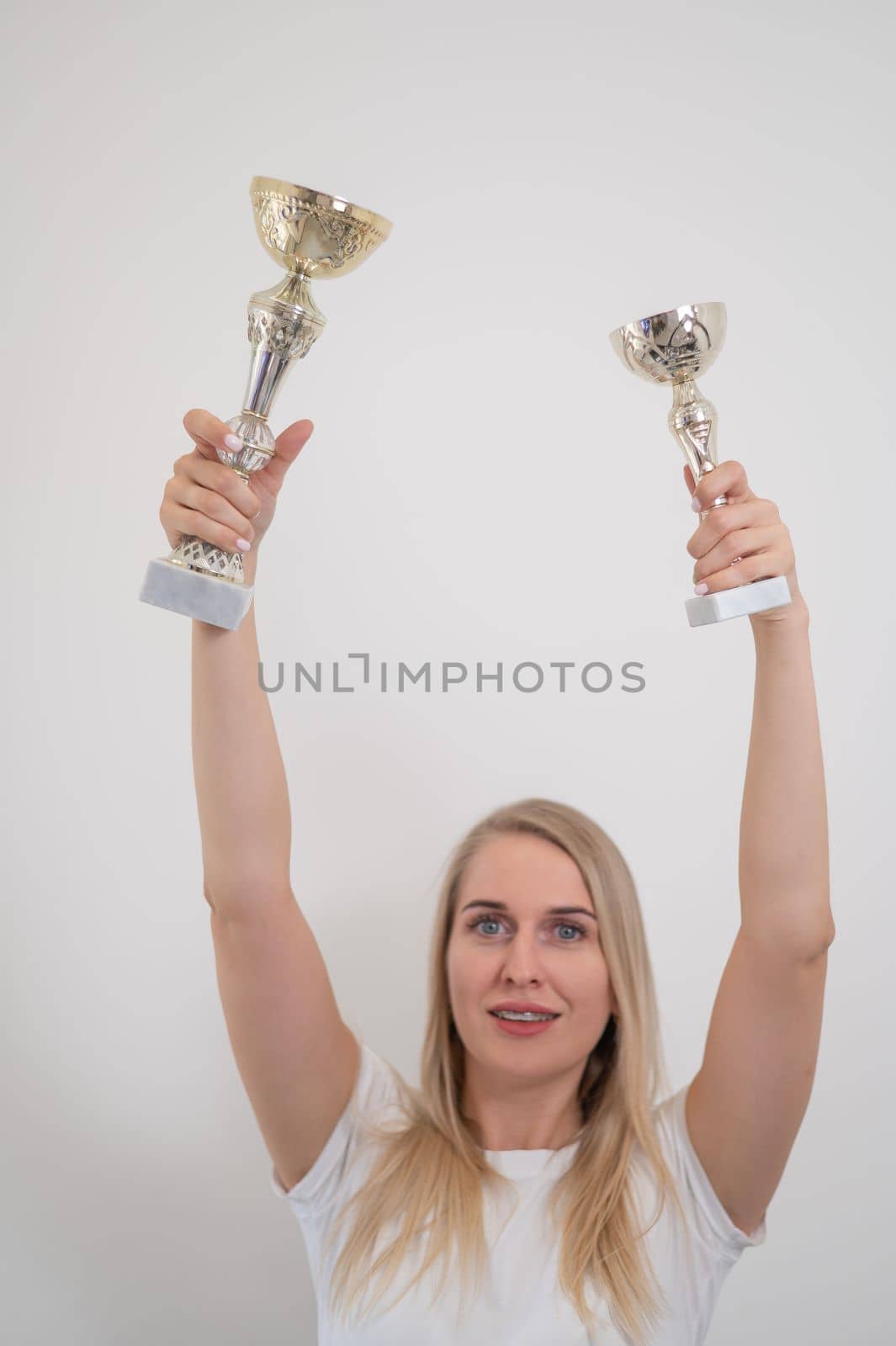 The blonde smiles and holds two golden trophy cup against the background of a white wall. by mrwed54