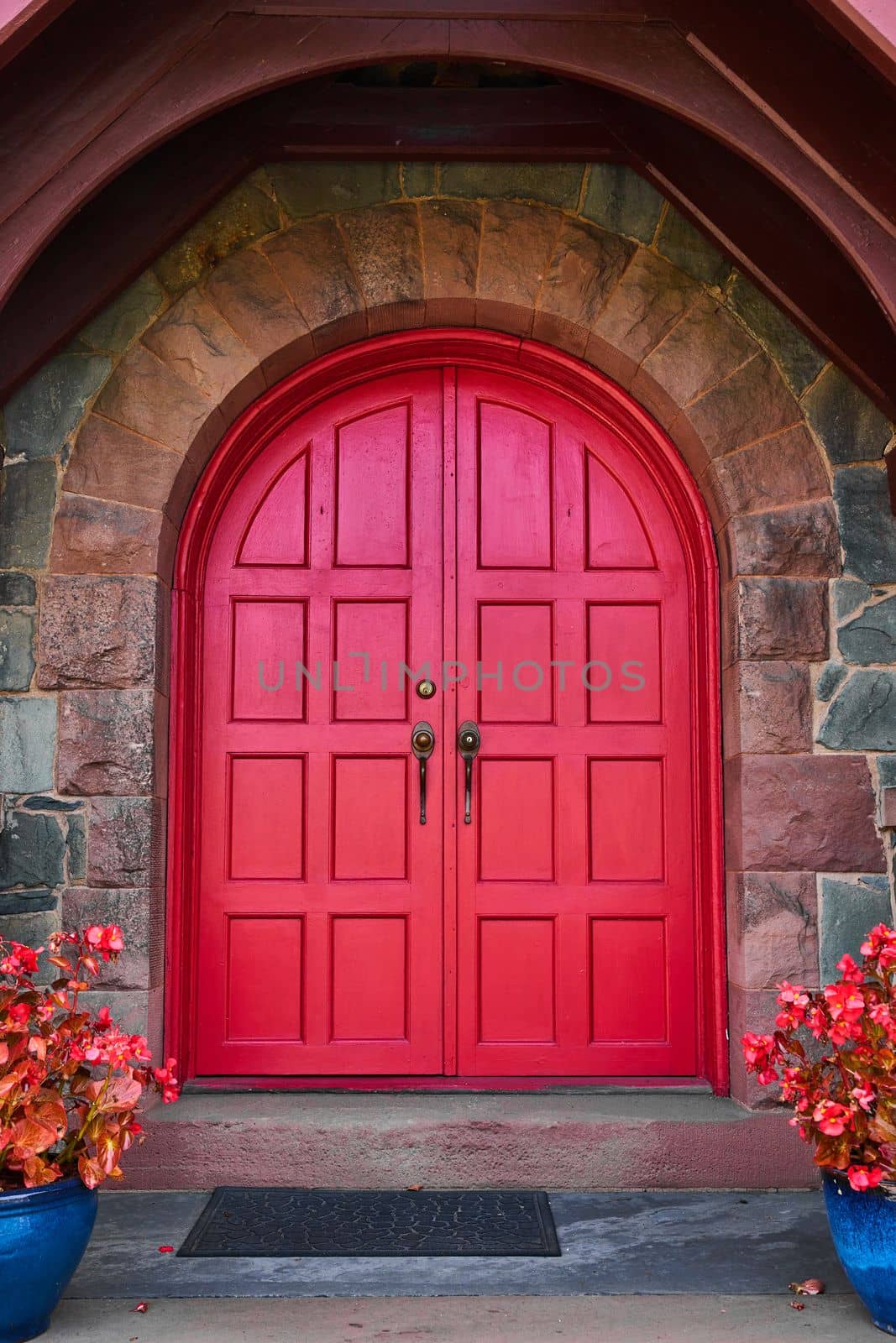 Image of Stunning vibrant red arched double doors with stone arch and pair of red plants in blue pots