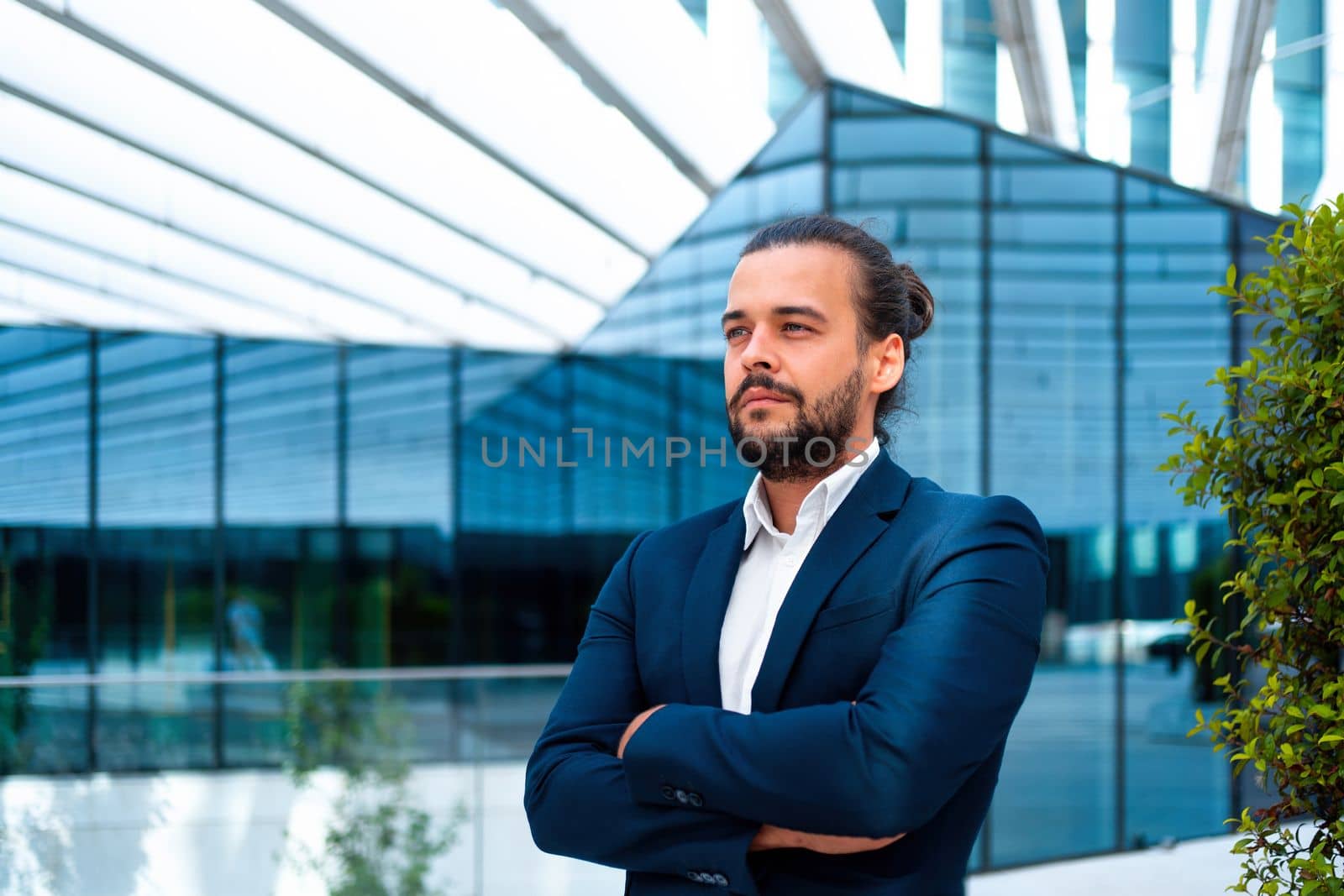 Confidence successful businessman or worker in suit with beard standing in front of office glass building arm crossed. Hispanic male business person portrait looking away. Free space