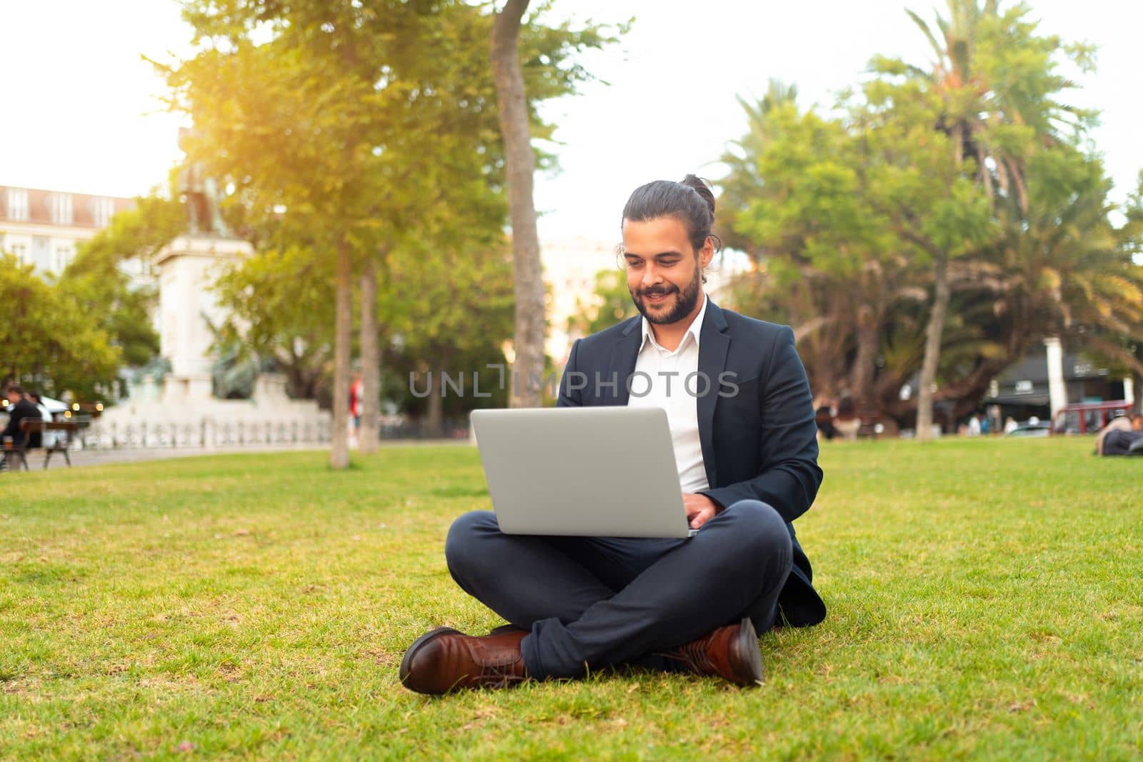 Handsome hispanic male businessman sitting lotus position in public park use laptop. Remote work concept. Stylish european man in suit relax in park summer day working on computer. Technology.