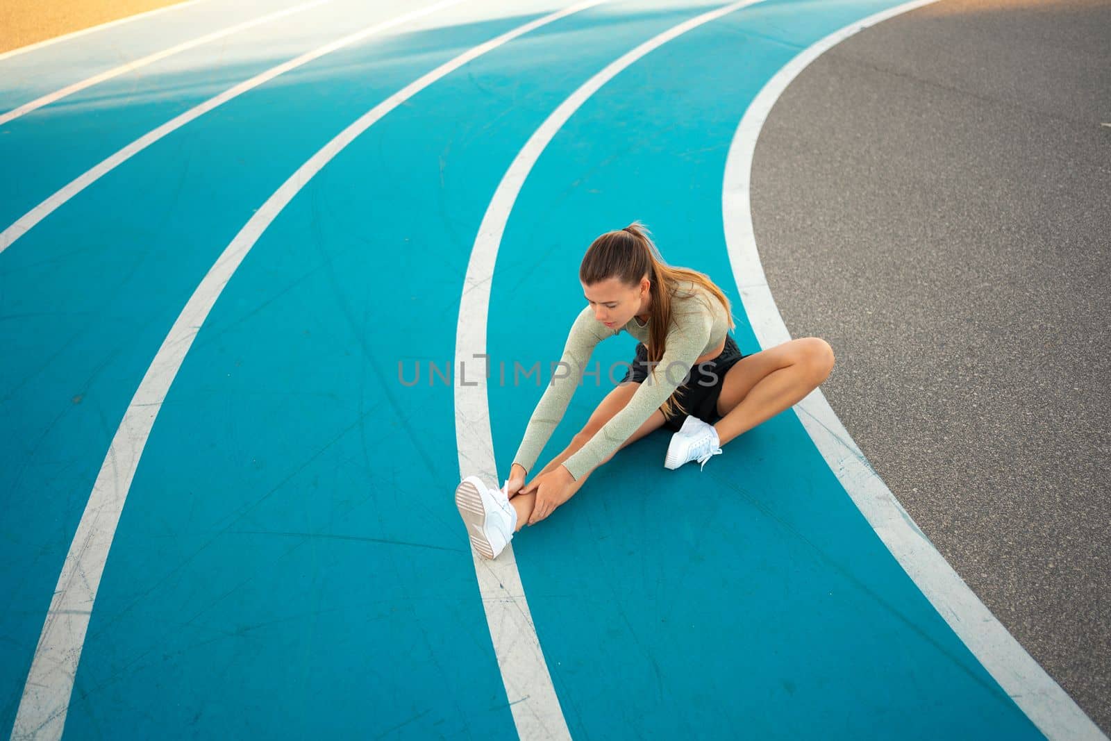 Sportswoman sitting and stretching on blue track field. Woman runner stretching leg muscles touching shoes. Warming up, Female runner doing stretching exercises sitting on running track. Free space