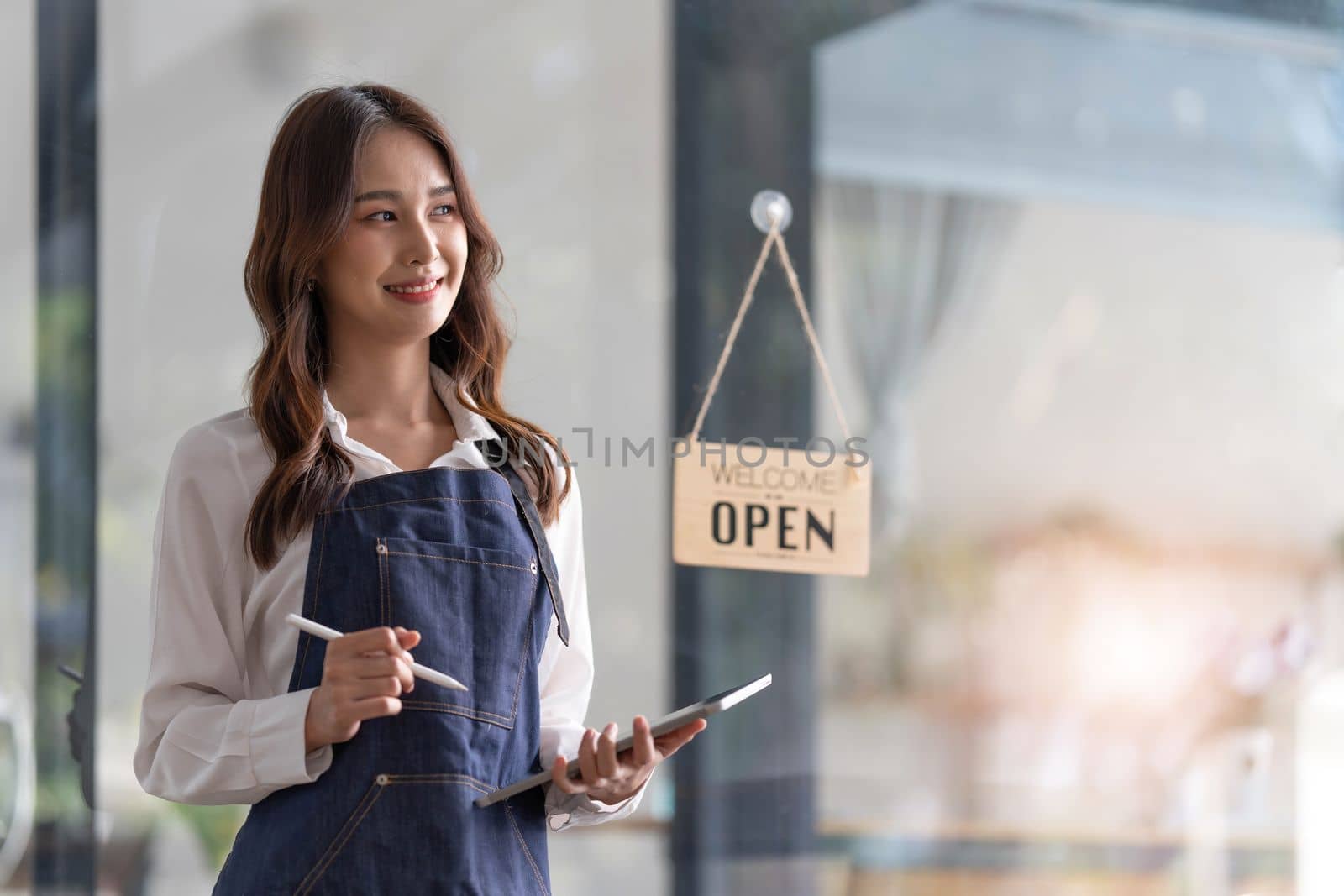 Beautiful asian young barista woman in apron holding tablet and standing in front of the door of cafe with open sign board. Business owner startup concept...