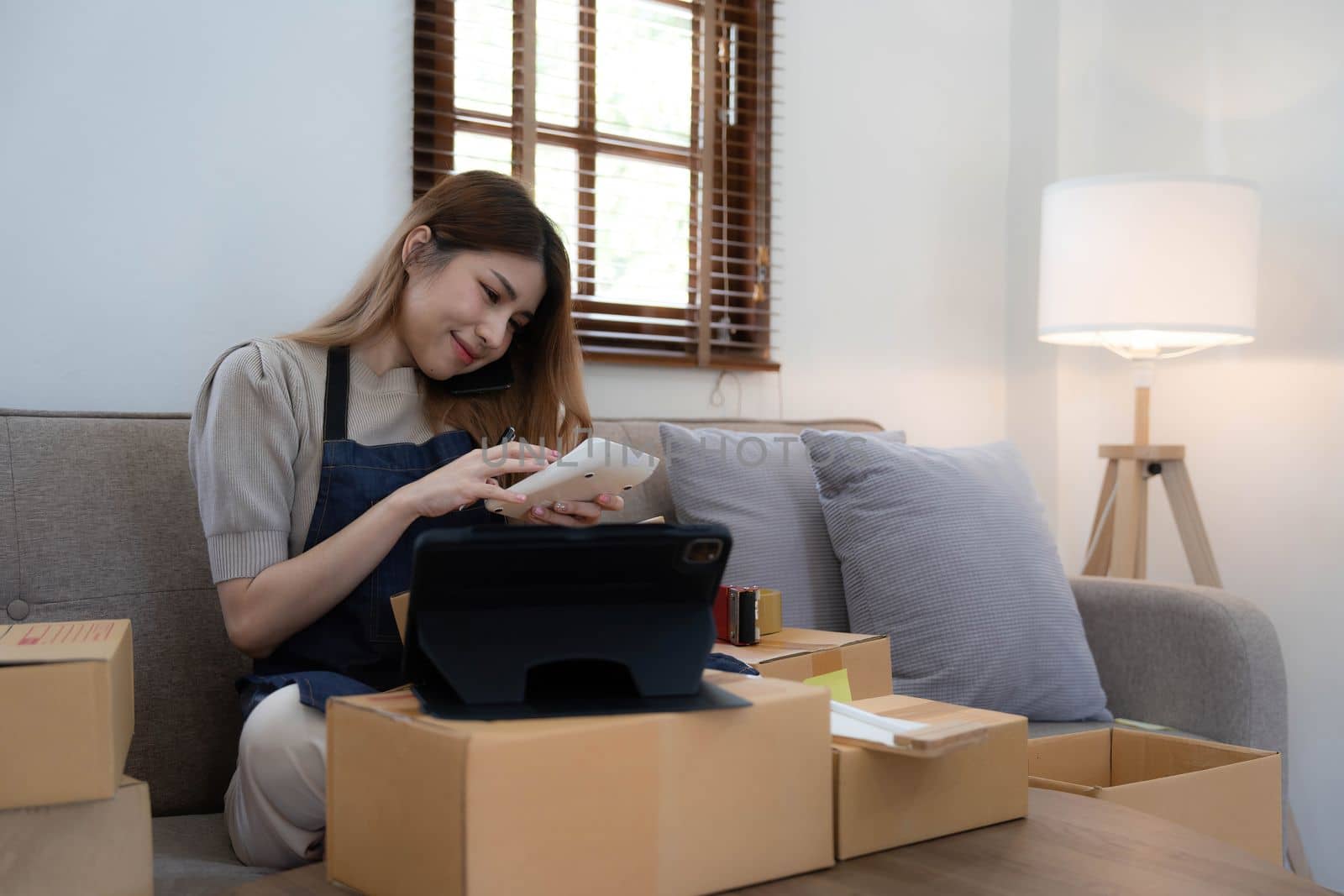 Startup SME small business entrepreneur woman using phone call receive from customer checking product on stock at home office, online marketing packaging delivery box