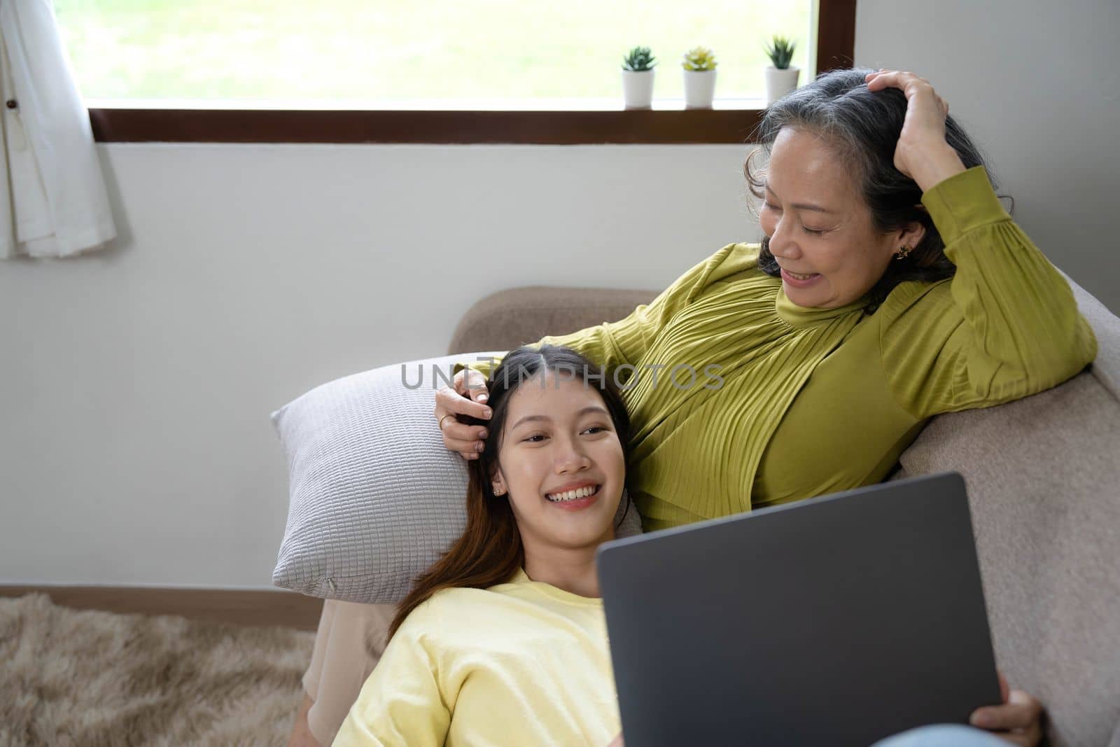 Happy adult granddaughter and senior grandmother having fun enjoying talk sit on sofa in modern living room, smiling old mother hugging young grown daughter bonding chatting relaxing at home together.