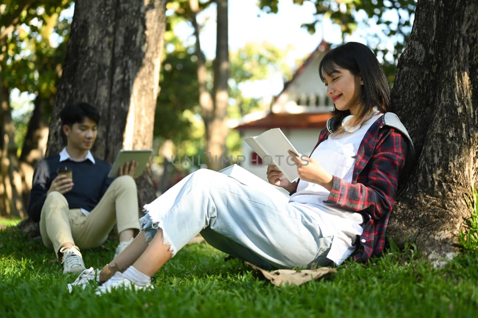 Smiling young woman sitting on lawn with friends and reading book. Education, learning, technology and lifestyle concept.