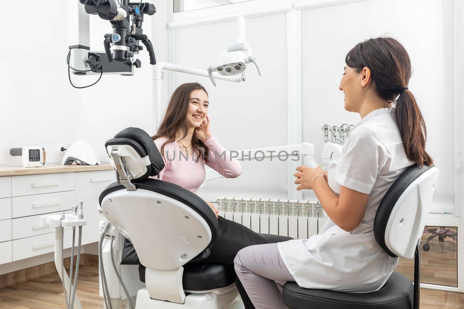 Female dentist talking to a young patient during appointment in modern dental clinic before teeth treatment by Mariakray