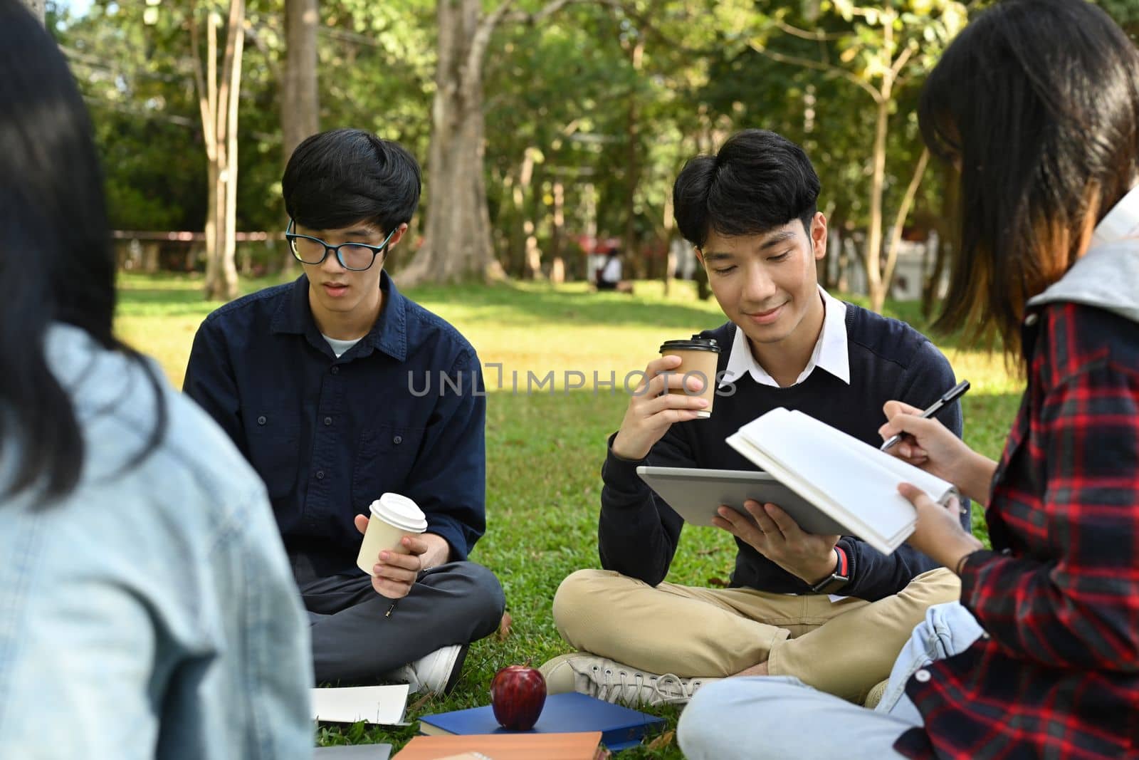 Group of university students talking with and reading book while sitting together on green grass in campus.