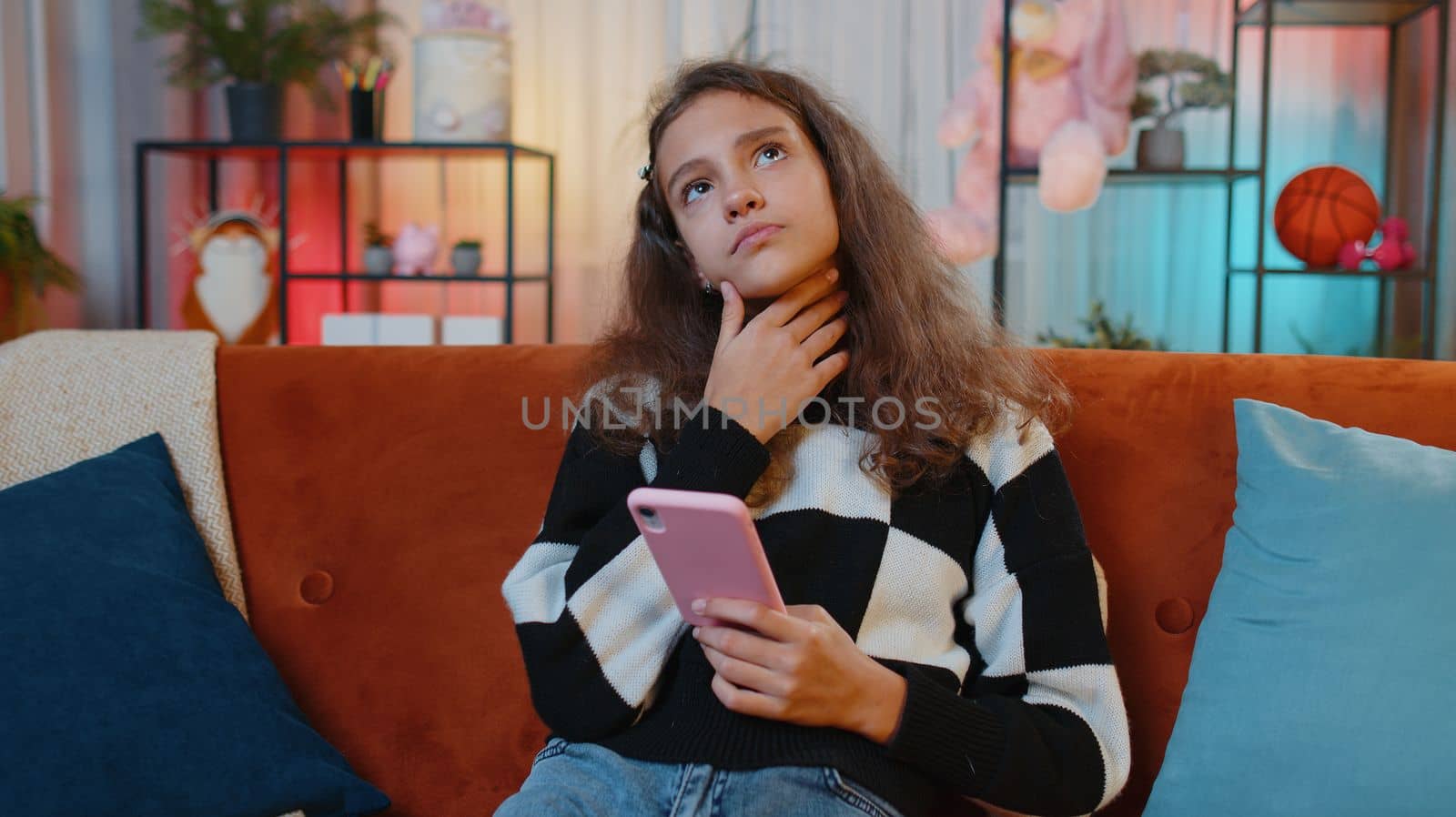 Preteen girl texting share messages content on smartphone social media applications online watching relax movie, news. Young child, kid at home in night evening living play room sitting on orange sofa