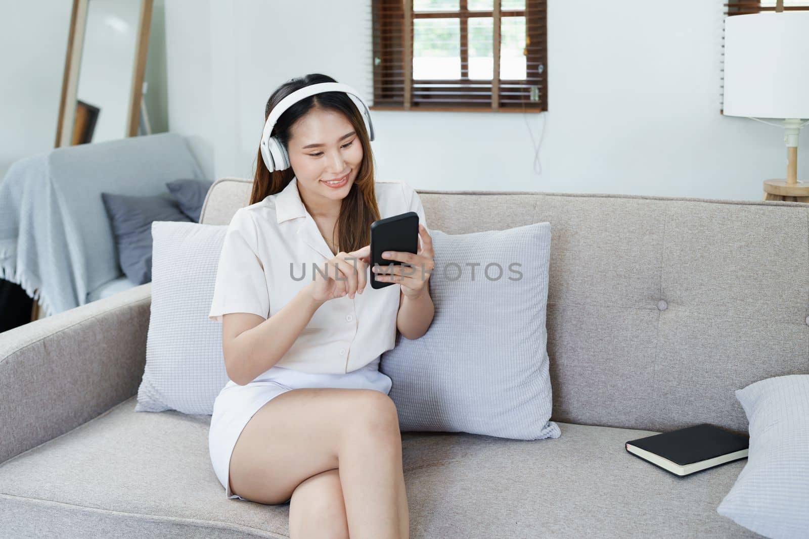 Portrait of asian woman using smart phone mobile and headphones relaxing on sofa at home by Manastrong