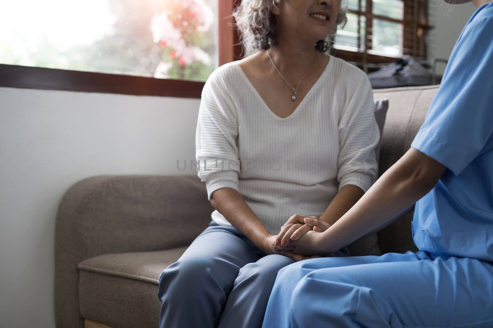 Happy patient is holding caregiver for a hand while spending time together. Elderly woman in nursing home and nurse...