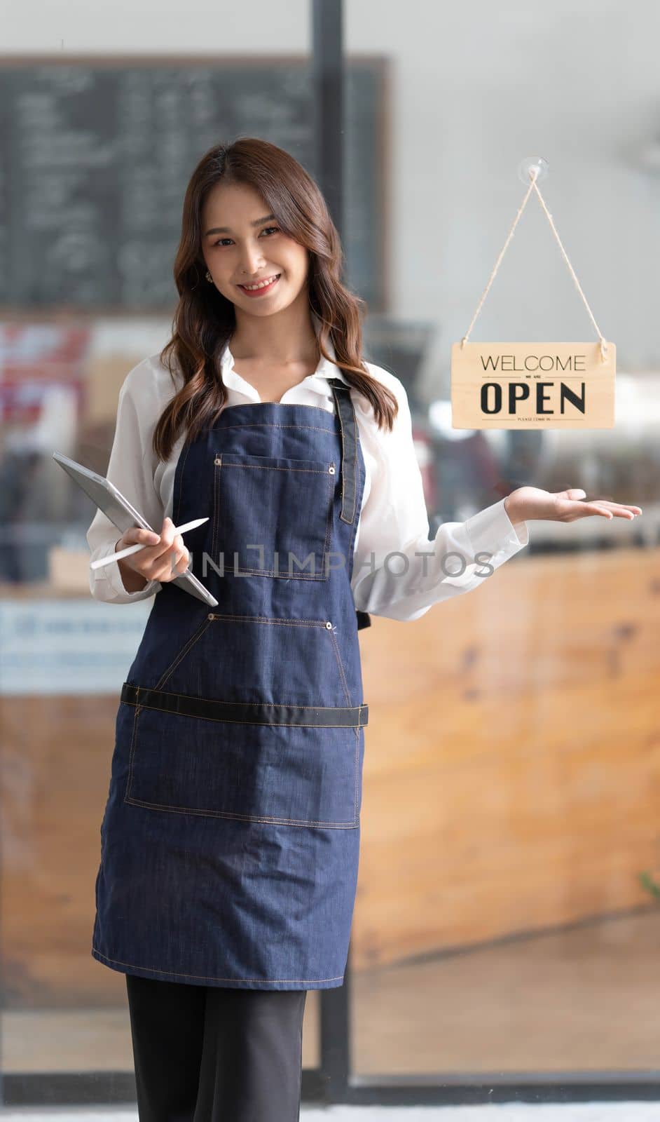 Beautiful asian young barista woman in apron holding tablet and standing in front of the door of cafe with open sign board. Business owner startup concept. by wichayada