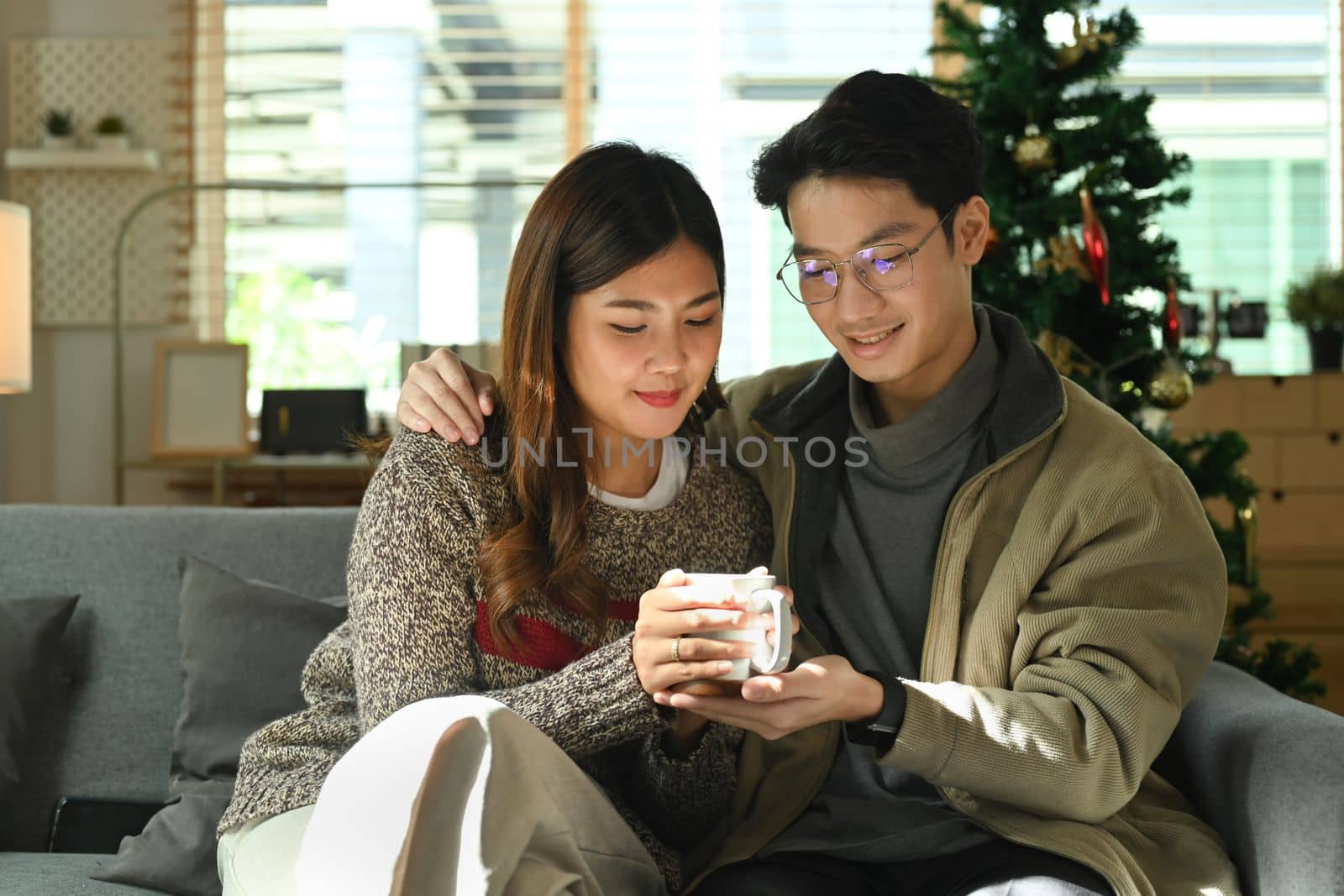 Romantic asian couple relaxing on comfortable couch, drinking hot chocolate and enjoying Christmas morning at home by prathanchorruangsak