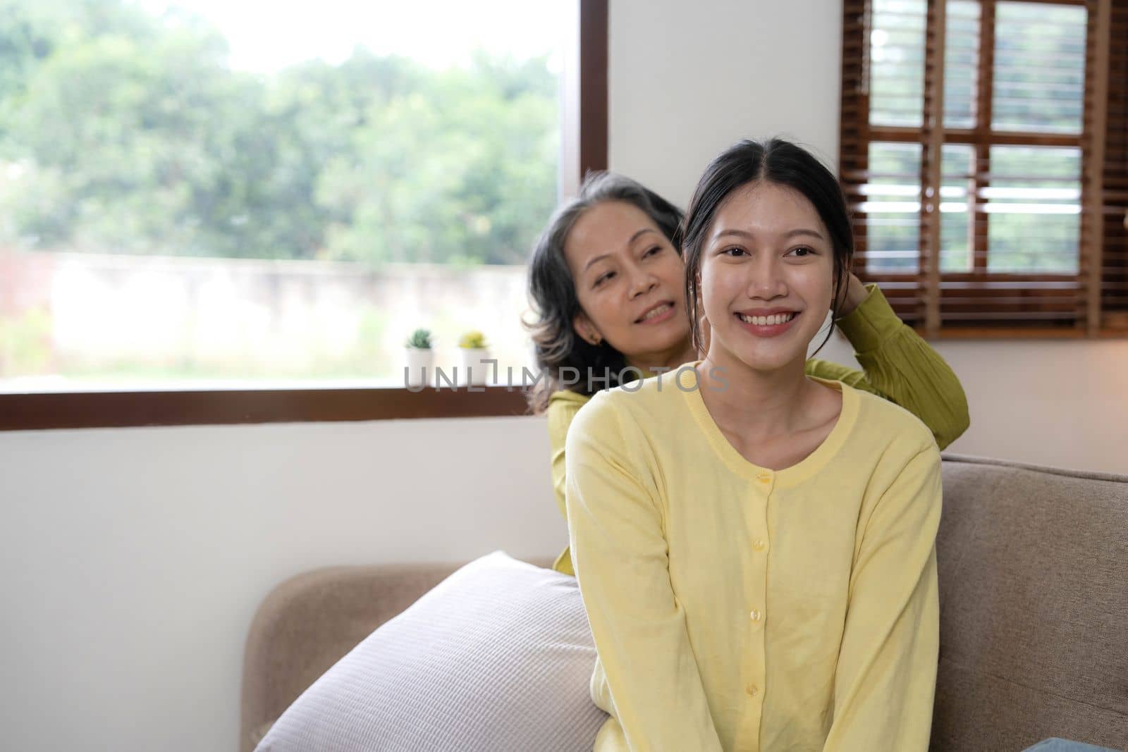 Happy adult granddaughter and senior grandmother having fun enjoying talk sit on sofa in modern living room, smiling old mother hugging young grown daughter bonding chatting relaxing at home together.