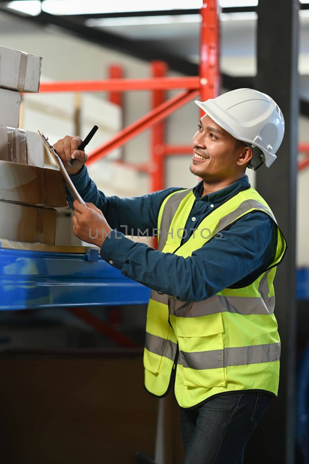 Smiling asian male worker using digital tablet and checking quantity of storage product on shelves full of packed boxes.