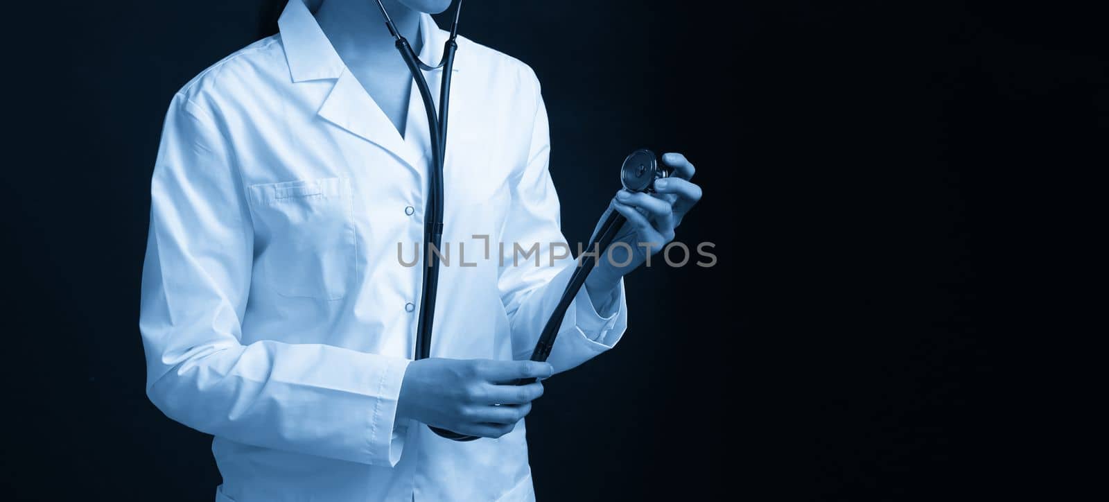 Cropped view of young female doctor holding stethoscope hanging on her neck, studio shot over black background with copy space by Mariakray