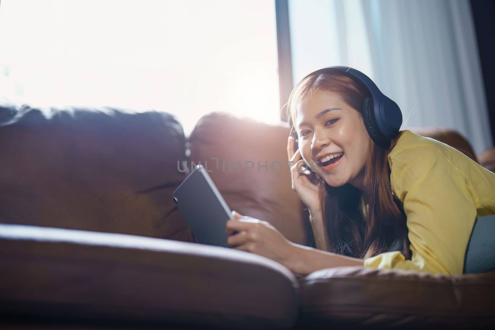 Portrait of asian woman using tablet and headphones relaxing on sofa at home by Manastrong