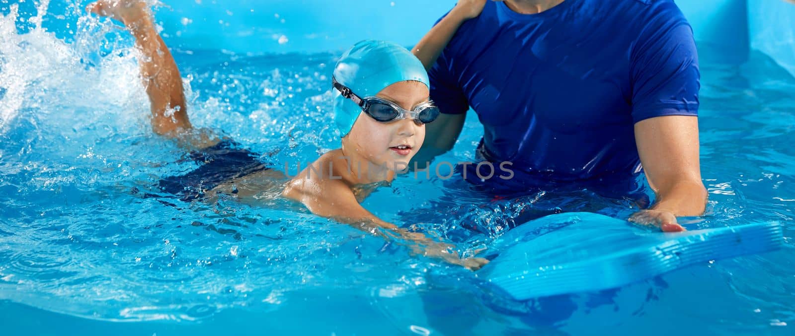 Trainer teaching little boy how to swim in indoor pool with pool floating board by Mariakray