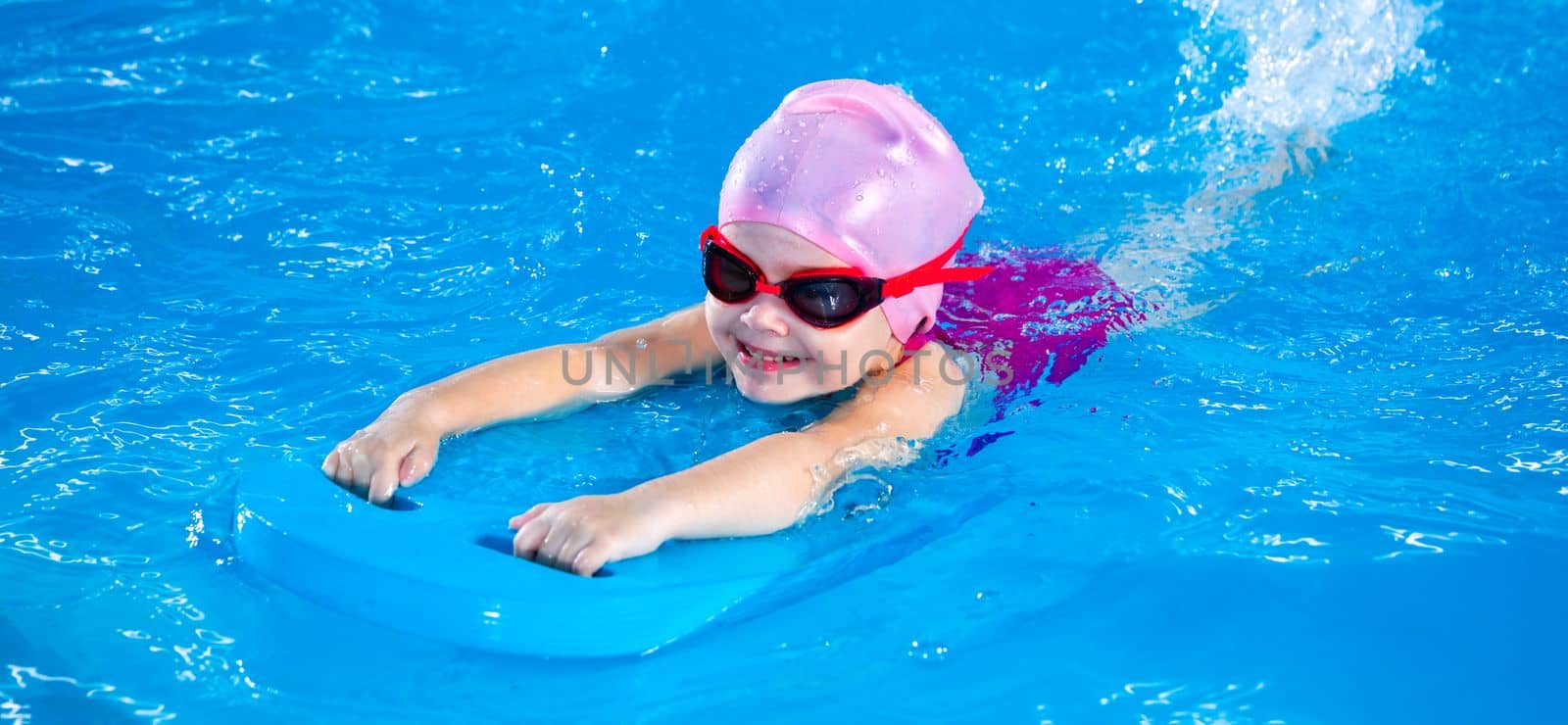 Smiling little girl learning to swim in indoor pool with flutter board during swimming class